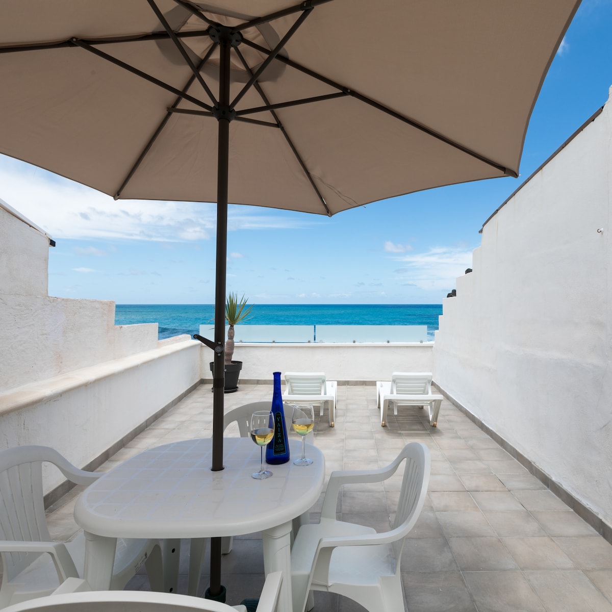 A spacious terrace is showcased, featuring a large umbrella providing shade over a table set with drinks and glasses. Two loungers are positioned nearby, all with a stunning view of the expansive ocean and bright blue sky in the background.