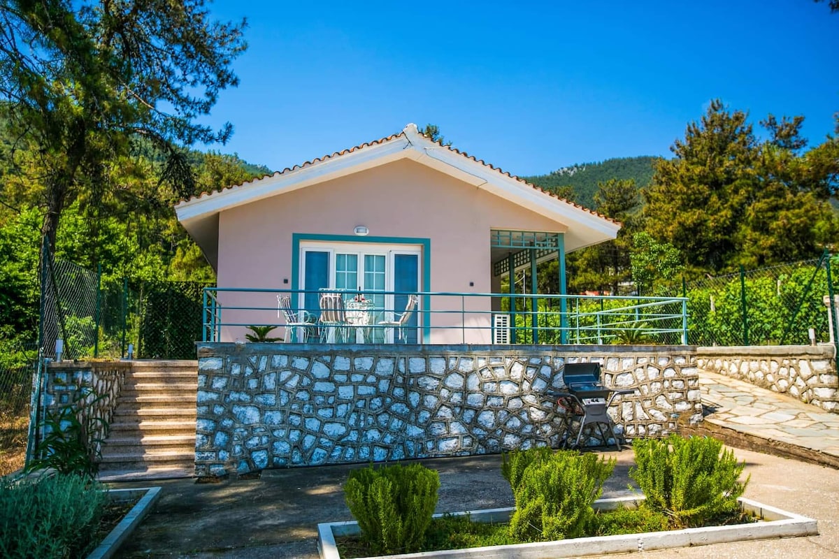 A renovated home is presented with a light pink exterior, featuring a stone wall and a staircase leading up to a spacious terrace. Outdoor seating is available, surrounded by greenery and trees, with a view of the scenic landscape.