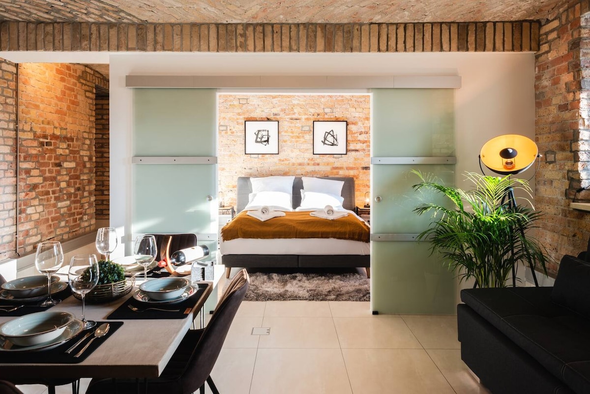 An open living area showcases a dining table set for four, accompanied by stylish tableware. A king-size bed is visible in the background, framed by glass sliding doors, against a backdrop of restored brick walls. A vibrant plant adds a touch of nature to the space.