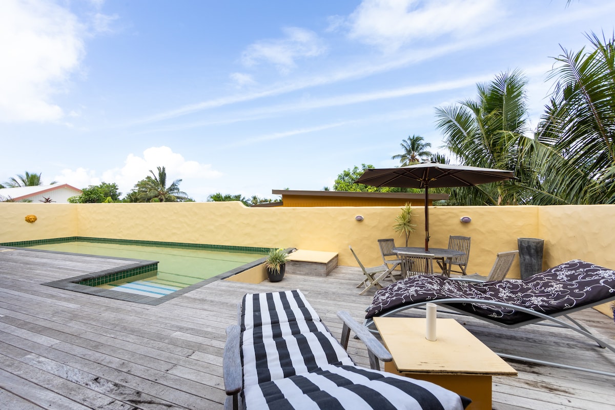 A sunlit terrace features a wooden deck surrounding a shared pool, with a lounge chair and a small table beside it. An umbrella provides shade over a dining area, complemented by tropical palm trees in the background and a yellow wall that creates a warm atmosphere.