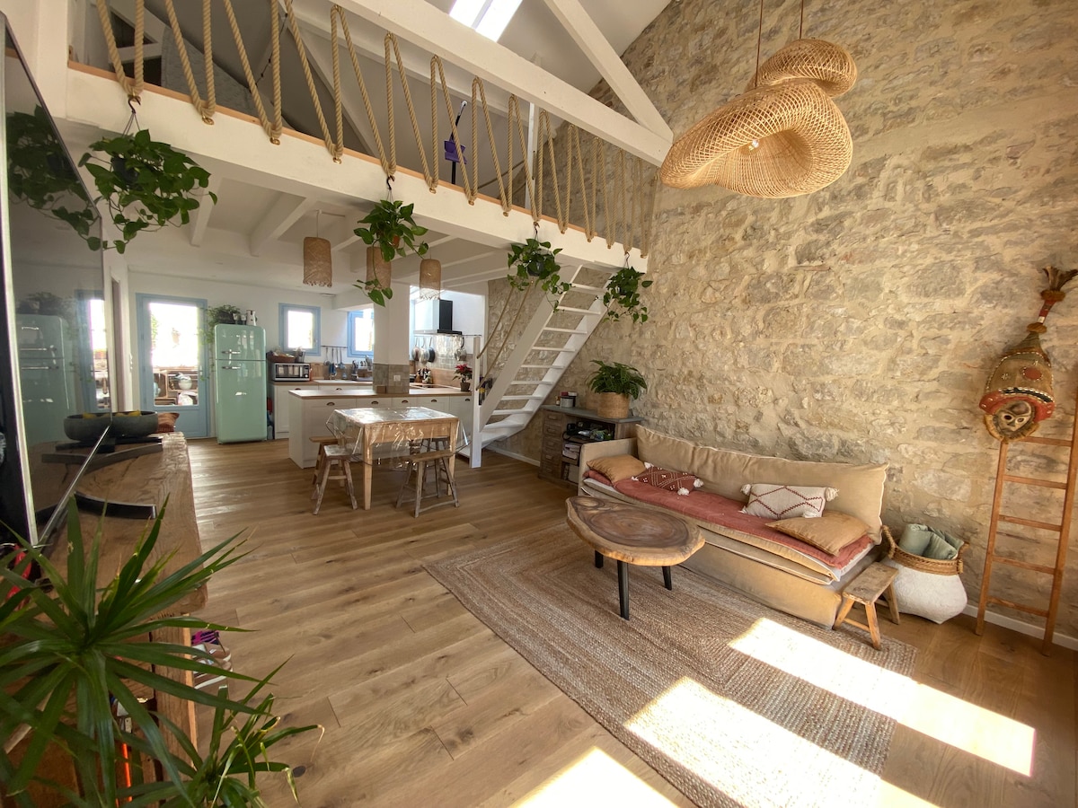 A spacious living area features a high ceiling with exposed beams and natural light flooding through large windows. A comfortable sofa is arranged with a low table on a woven rug, alongside greenery and decorative elements. The nearby open kitchen includes modern appliances and a dining area.