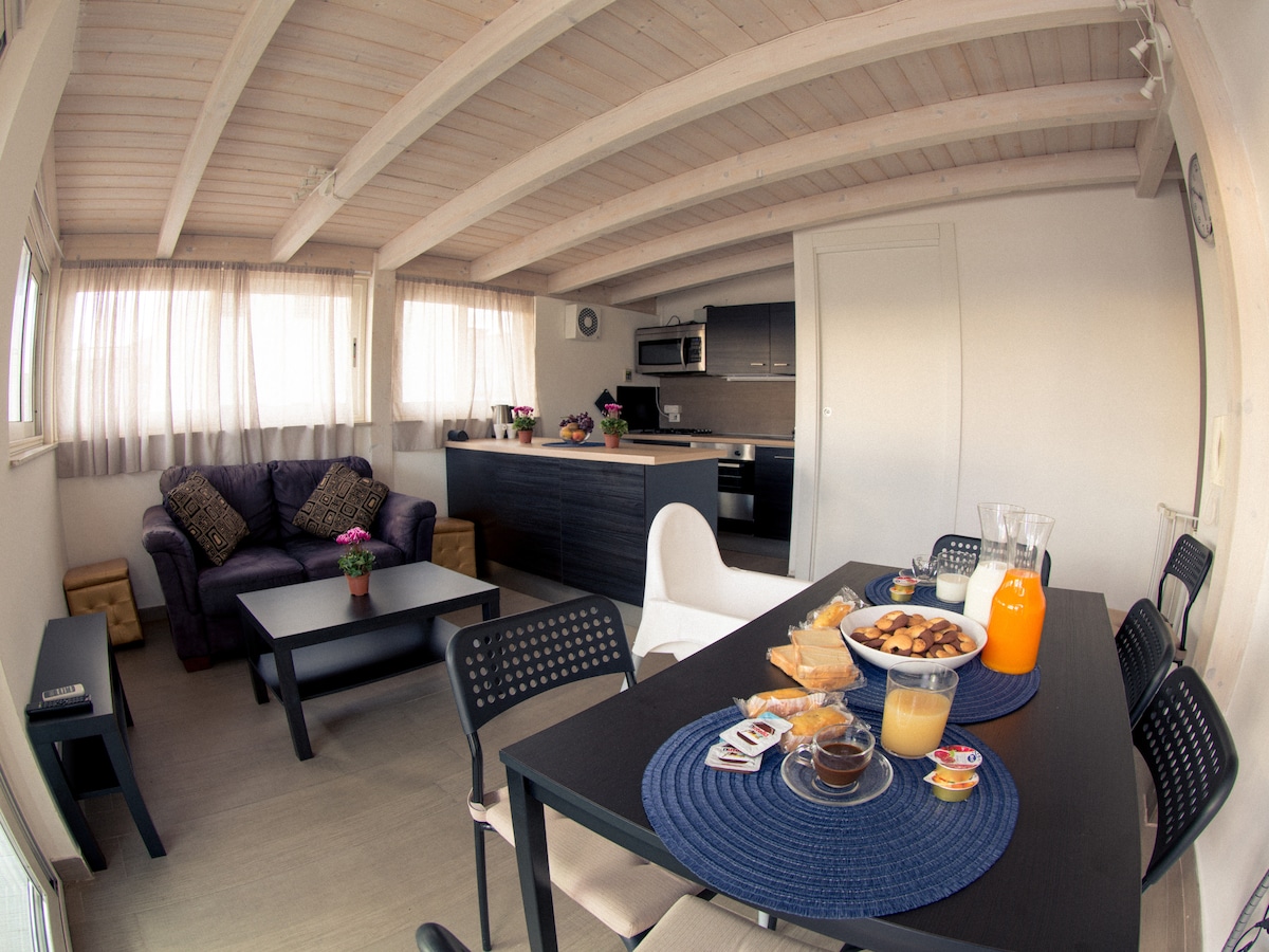 An open living area features a purple sofa adorned with decorative cushions. A dining table is set with breakfast items, including juice and pastries. The space is illuminated by natural light through large windows, and modern kitchen appliances are visible in the background.