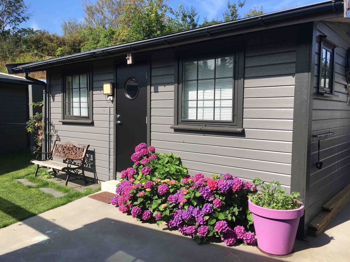 The image displays a small, gray exterior of a building, featuring a round window and a black door. Vibrant hydrangeas in purple bloom are planted along the pathway and next to a large purple pot. A wooden bench is positioned on the side.