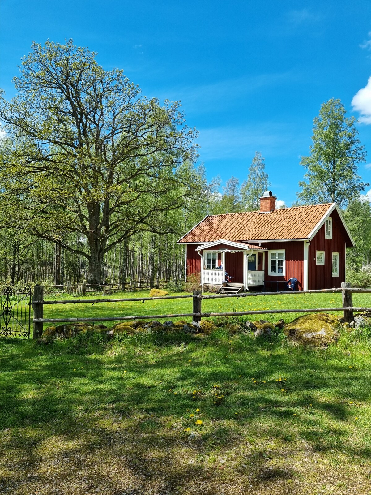 A red cottage with white trim sits surrounded by lush greenery and tall trees under a clear blue sky. A porch extends from the front, offering a welcoming entryway. A wooden fence delineates the property, enhancing the natural setting.