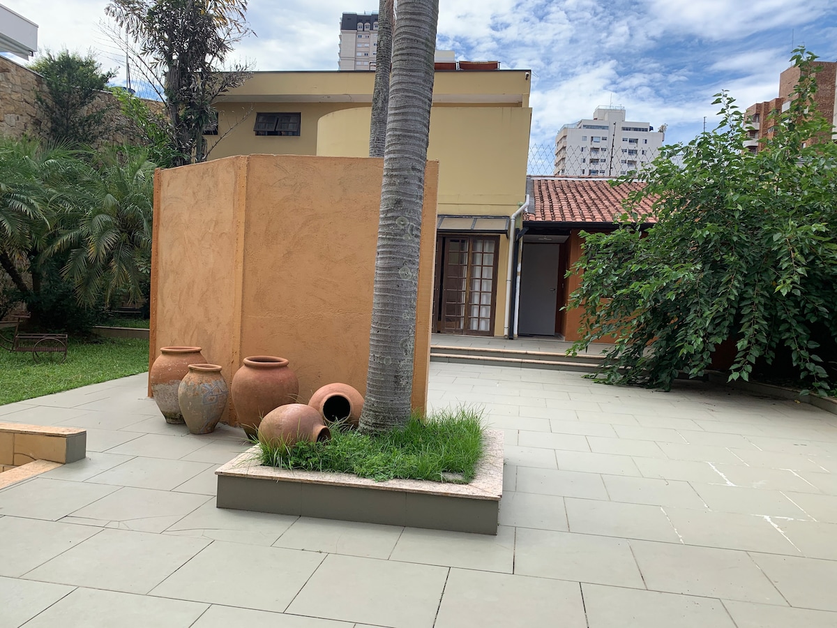 An outdoor patio area features light-colored stone flooring and a lush green lawn. A tall palm tree stands beside a terracotta urns arrangement. The background displays a building with a red-tiled roof and large glass doors leading to an interior space.