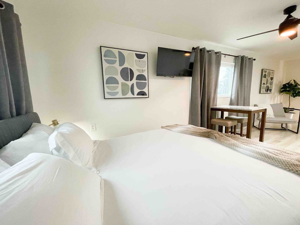 A studio apartment features a queen bed with crisp white bedding, positioned near a window. A wall-mounted smart TV is visible on one side, alongside a small dining table with stools. Light gray curtains frame the window, enhancing the overall brightness of the space.