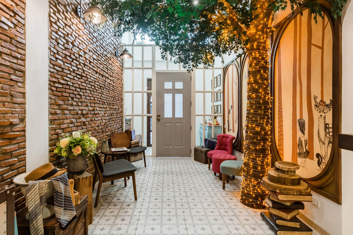 The entryway features a blend of exposed brick walls and artistic tree-like decorations illuminated by warm string lights. A variety of seating options, including vintage chairs, is arranged around the space, while a patterned floor adds a distinctive touch.