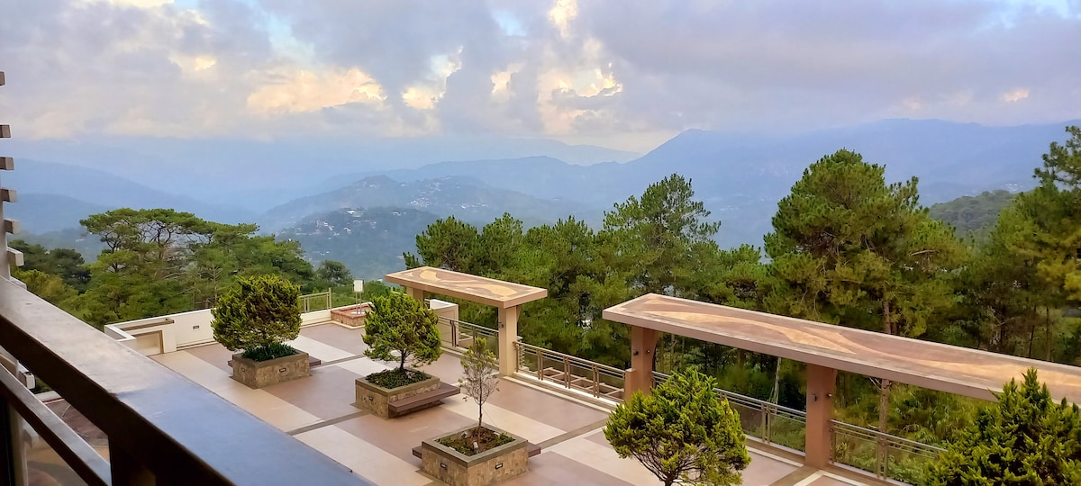 A spacious outdoor terrace is framed by tall trees, providing stunning views of distant mountains. Two large stone tables are positioned under shaded structures, surrounded by carefully maintained greenery. Soft clouds drift in the sky above, enhancing the serene ambiance of the space.