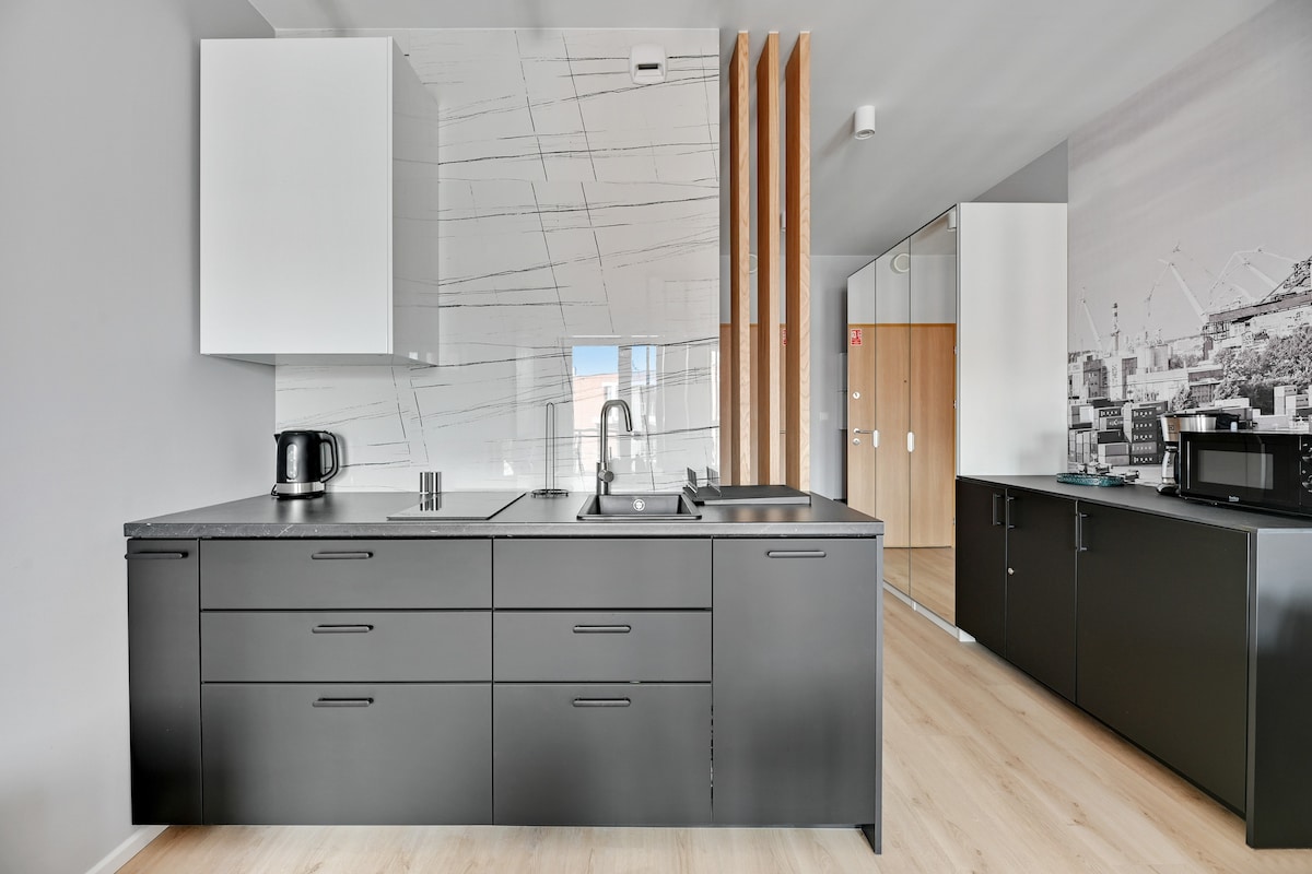 A modern kitchen area features sleek black cabinets and a stylish countertop. Essential appliances, including a kettle and a microwave, are positioned conveniently. Natural light illuminates the space through a window, complementing the minimalist design with a light wooden floor.