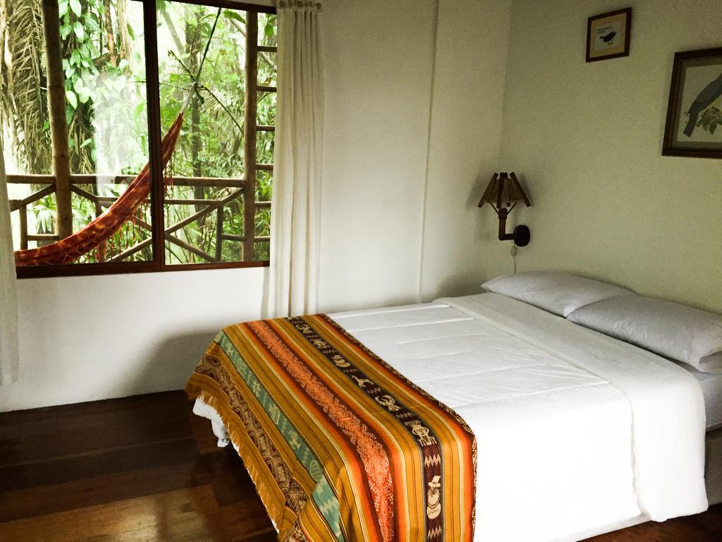 A simple and inviting sleeping area is presented, featuring a spacious bed with white linens. A colorful woven blanket adds a warm touch. Natural light filters through a large window, revealing lush green foliage outside. A hammock is visible, enhancing the relaxing atmosphere.