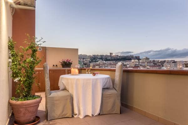 A spacious terrace features a round dining table draped with a white tablecloth, surrounded by two cushioned chairs. Potted plants add greenery, while a scenic view of the city skyline, including historical buildings, is visible in the background under a serene sky.