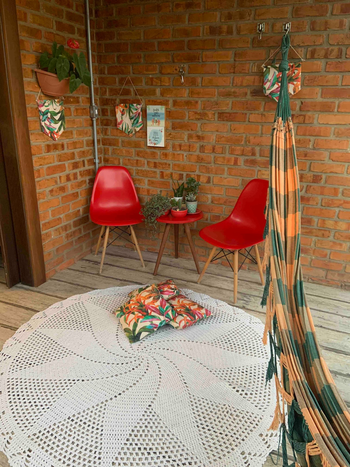 A cozy outdoor seating area features two vibrant red chairs positioned around a small table, adorned with potted plants. A white crochet rug lies underfoot, complemented by decorative cushions. Hammocks hang nearby, inviting relaxation in this warm, rustic space.