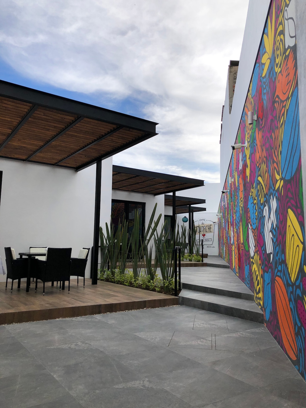 A vibrant mural adorns the white wall, showcasing a variety of colorful designs. The pathway leads to a patio space featuring a dining set with four black chairs and a table. Lush greenery, including tall plants, adds to the outdoor ambiance.