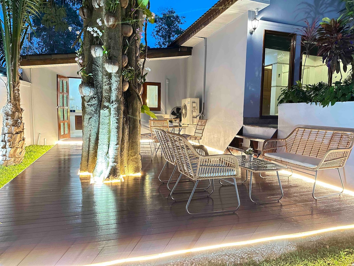 An outdoor seating area features several woven chairs surrounding a glass table. Soft lighting highlights the space, with a mature tree providing partial shade. The area is bordered by lush greenery, contributing to a peaceful atmosphere.