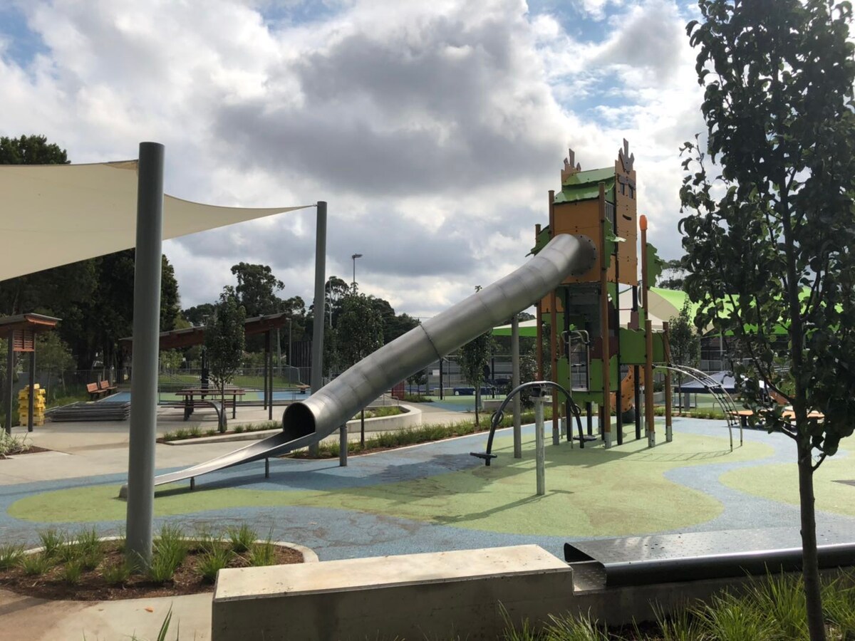 A modern playground features a large metallic slide leading from a multi-level structure. The play area is surrounded by soft landscaping, shaded by fabric canopies. Benches and open spaces are visible in the background, with trees providing greenery in the vicinity.
