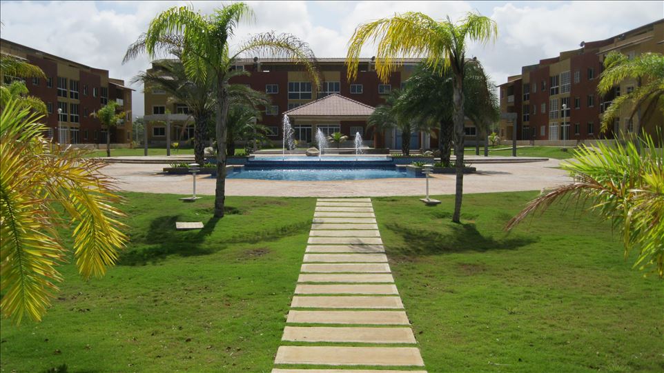 A landscaped area features a pathway leading towards a central fountain surrounded by greenery. Tropical palm trees are positioned around the spacious lawn, with an inviting pool located in the backdrop of the colorful apartment buildings.