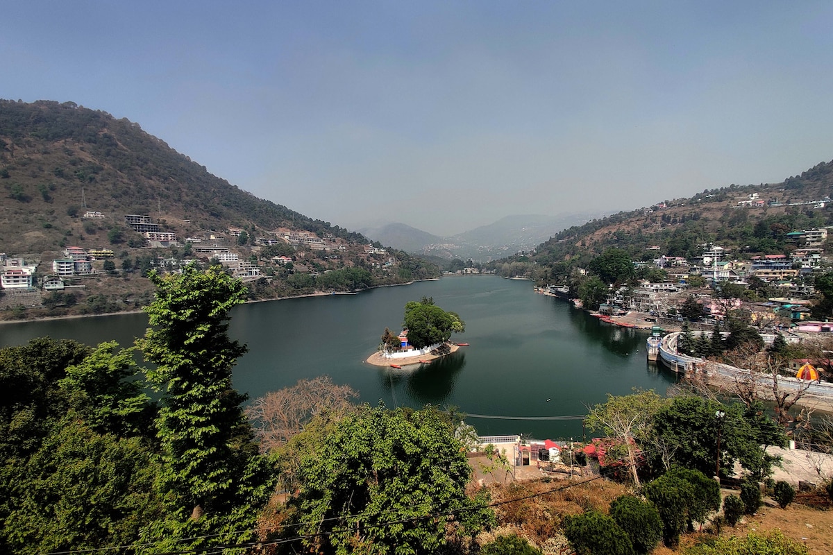 A serene view of Bhimtal Lake is captured, framed by lush green trees and rolling hills. An island with a small structure is visible in the center of the lake, while scattered houses dot the surrounding terrain, creating a harmonious blend of nature and settlement.