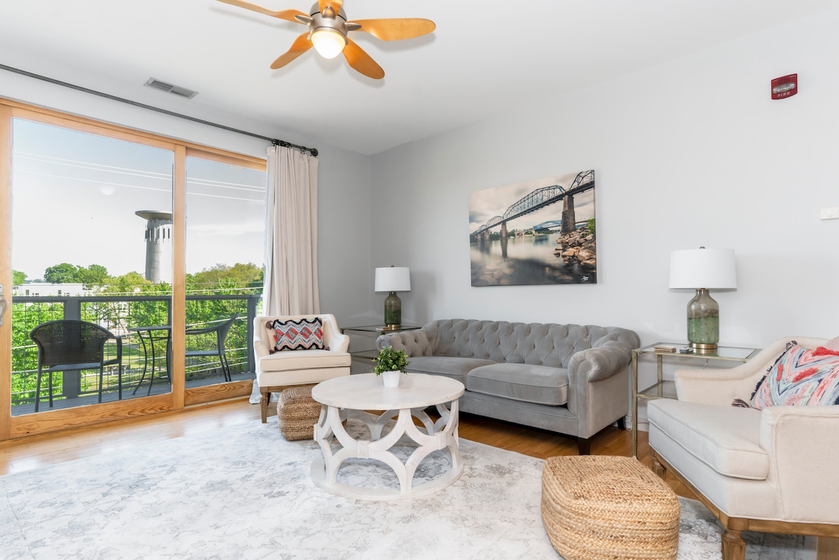 A comfortable living area features a gray tufted sofa and a round coffee table, surrounded by natural light from sliding glass doors. A light-colored area rug lies beneath, complemented by a decorative chair and two lamps on side tables. A city view is visible through the windows.