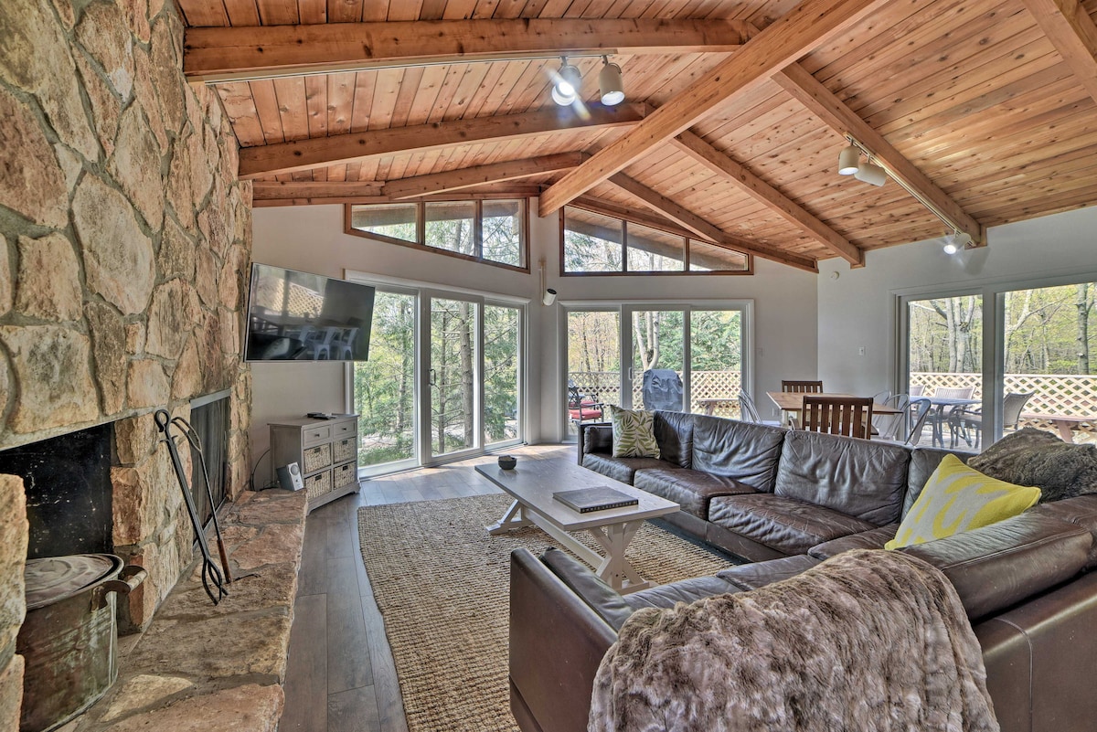 A spacious living area features a large leather sectional sofa, complemented by a cozy throw blanket. A stone fireplace serves as a focal point, with a mounted television above. Expansive glass doors allow natural light to illuminate the room, providing views of the surrounding woodlands.