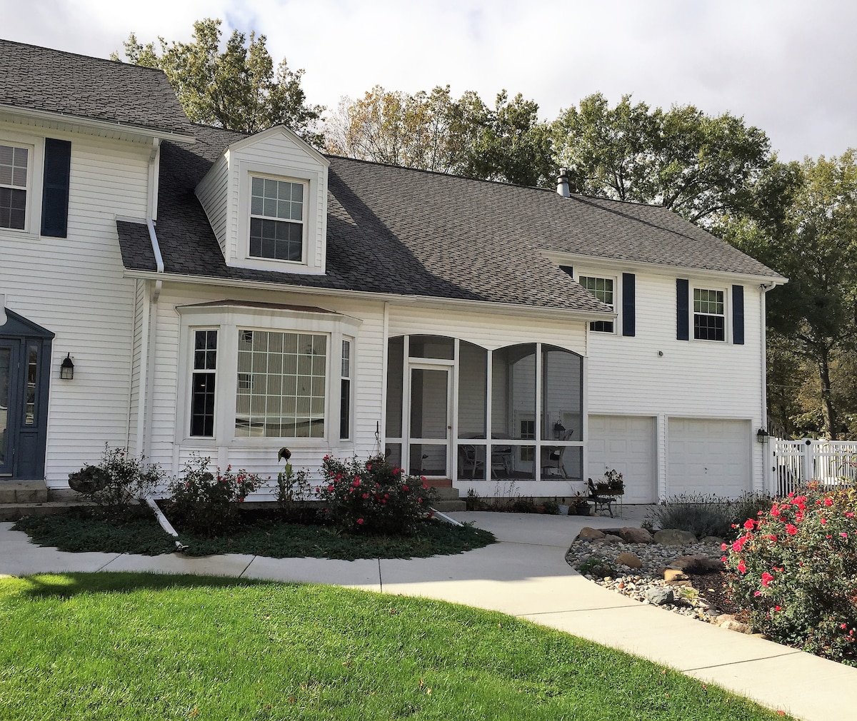 An inviting exterior view of a farmhouse showcases a front porch with a spacious screened area. Lush green grass and well-maintained flower beds frame the entrance. Large windows allow natural light to illuminate the interior, and a driveway leads toward the garage on the right.