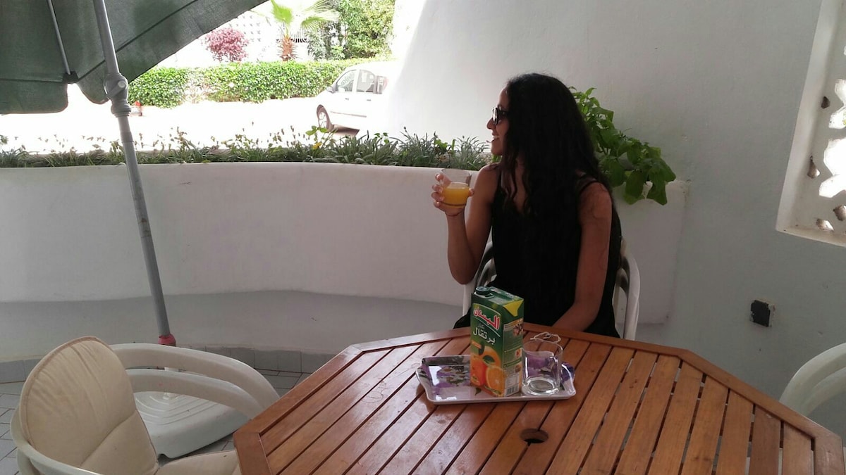 A cozy outdoor seating area features a wooden table surrounded by white chairs. A tray displays beverages and snacks, while lush greenery is visible in the background. A large umbrella provides shade, enhancing the inviting environment for relaxation.
