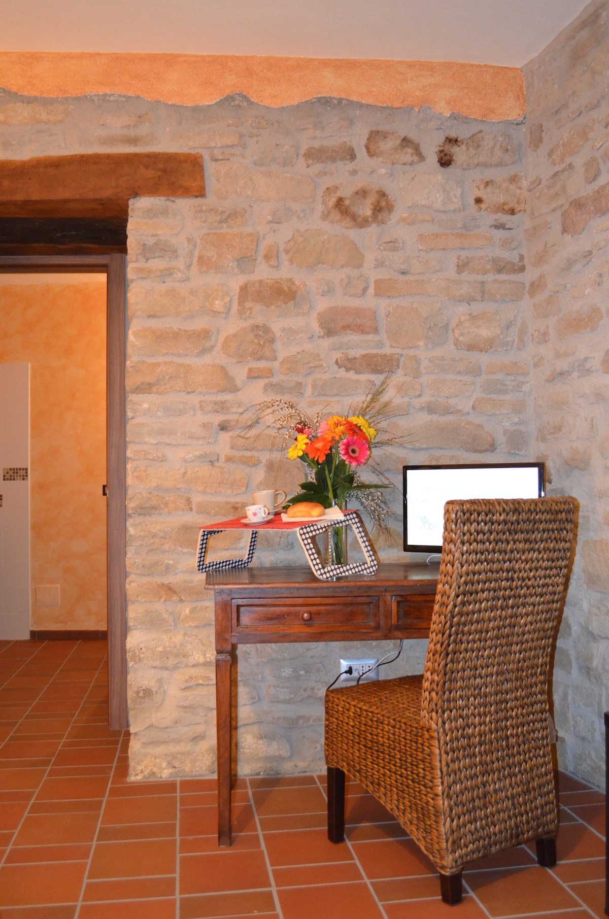 A cozy workspace is positioned against a natural stone wall, featuring a wooden desk with a computer and decorative frames. A chair with woven detailing complements the setup. Fresh flowers in a vase add a touch of color, enhancing the inviting environment.