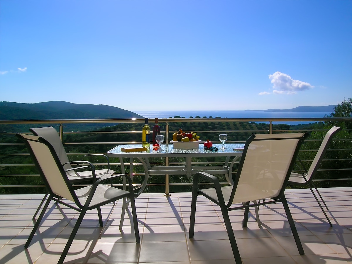 An outdoor dining area is displayed, featuring a glass table surrounded by four chairs. A bottle of wine and glasses are set on the table, accompanied by fresh fruit. The expansive view shows green hills and blue waters under a clear sky.