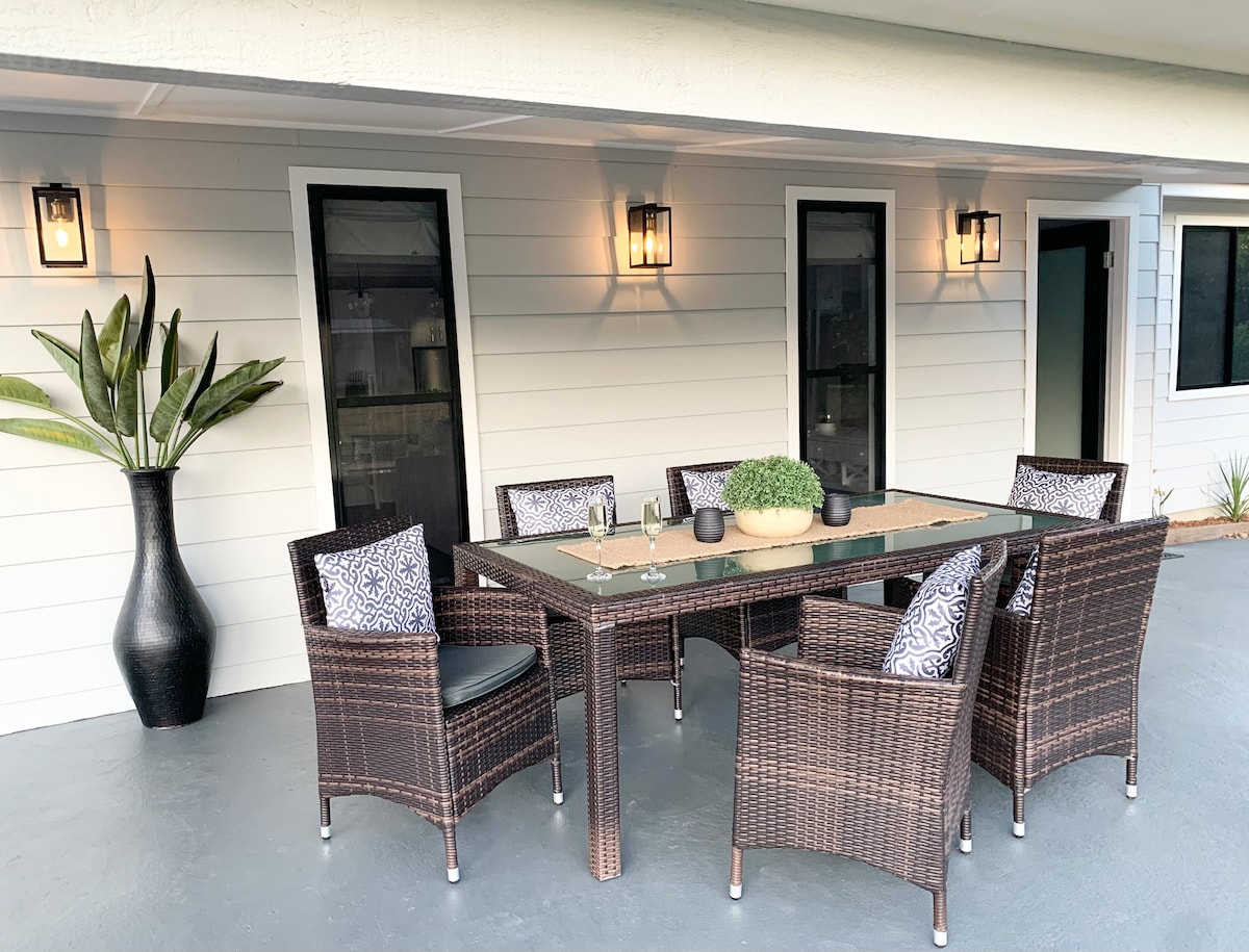 An outdoor dining area features a rectangular table surrounded by six woven chairs, each adorned with patterned cushions. A decorative vase with greenery adds a touch of nature. Soft lighting from mounted fixtures illuminates the space, enhancing a pleasant atmosphere for gatherings.