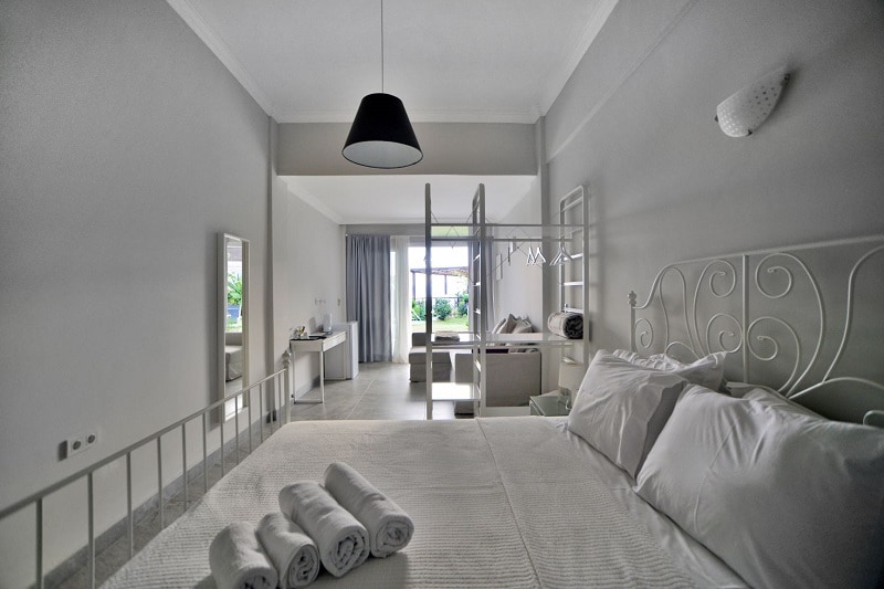 A bright sleeping area features a spacious bed with neatly rolled towels placed on top. Light-colored walls and furnishings create an airy feel. Large glass doors allow natural light and views of the outdoor space. A modern seating area is visible in the background.