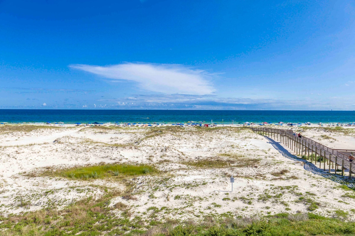 An unobstructed view of the beach is presented, showcasing fine white sand and the expansive turquoise ocean. A wooden boardwalk leads to the shoreline, while colorful beach umbrellas dot the landscape under a clear blue sky.