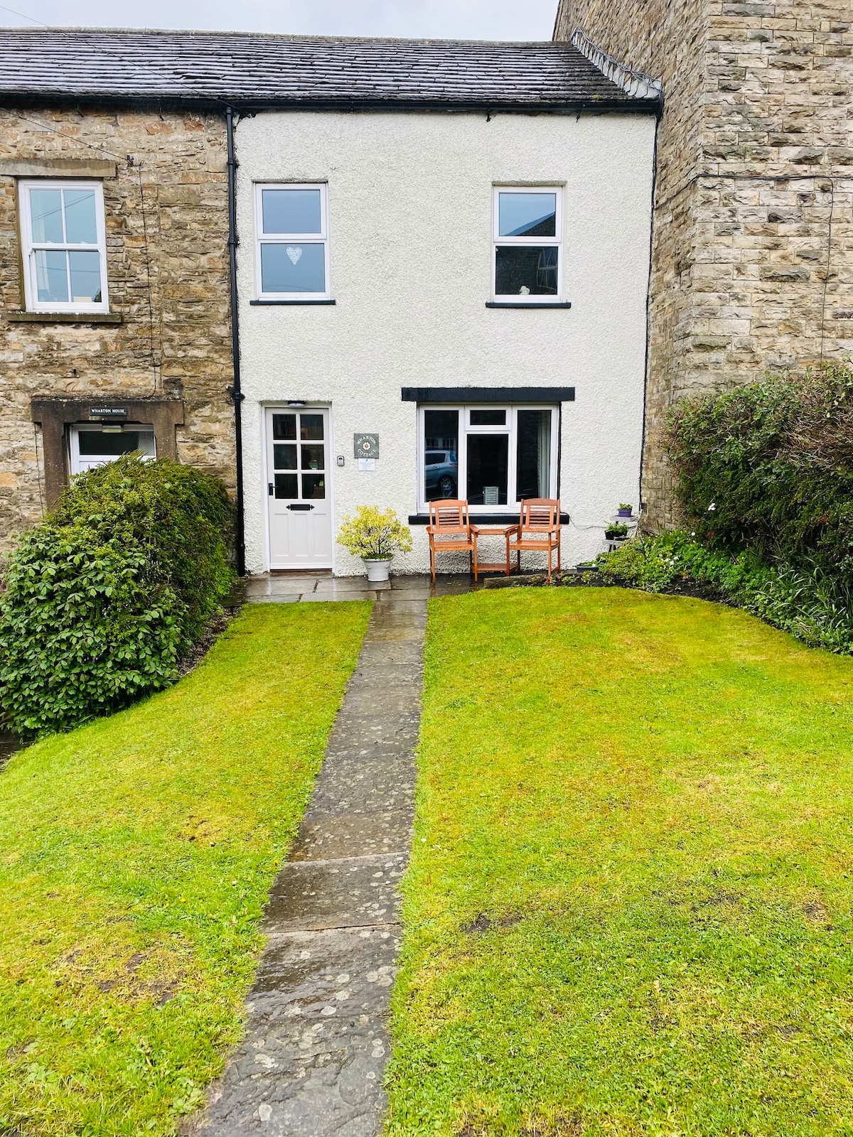 The front of the cottage features a well-maintained lawn garden with a stone pathway leading to the entrance. Two wooden chairs are positioned beside a small table, offering a welcoming spot to relax outdoors. The exterior is a combination of stone and white render, creating a quaint appearance.