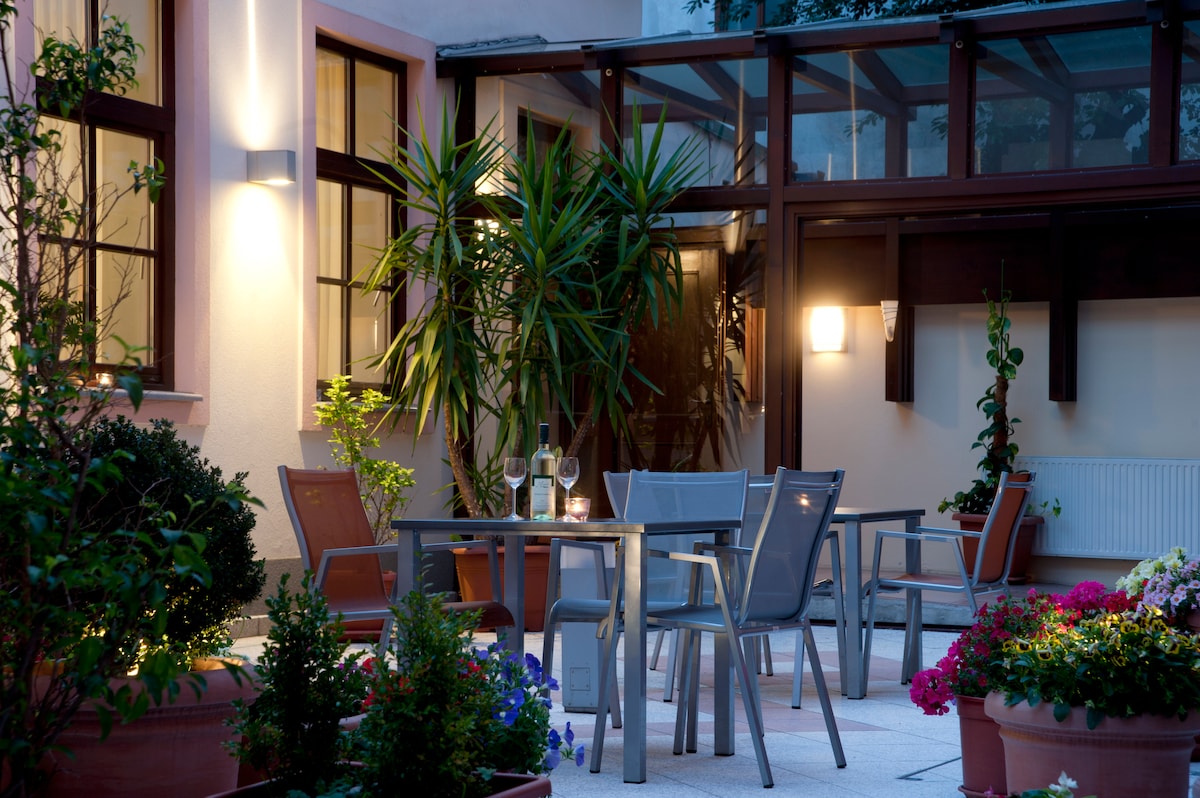 A tranquil courtyard features sleek chairs and a table, surrounded by lush greenery and colorful flower pots. Soft lighting enhances the evening ambiance, creating an inviting space for relaxation. The glass enclosure adds to the charm, providing a seamless connection to nature.