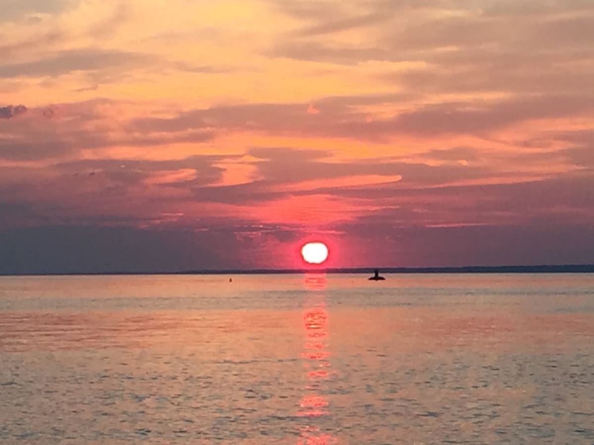 A serene sunset is depicted over calm waters, reflecting shades of orange, pink, and purple. The sun hovers just above the horizon, with a faint silhouette of a lighthouse visible in the distance, enhancing the tranquil atmosphere.