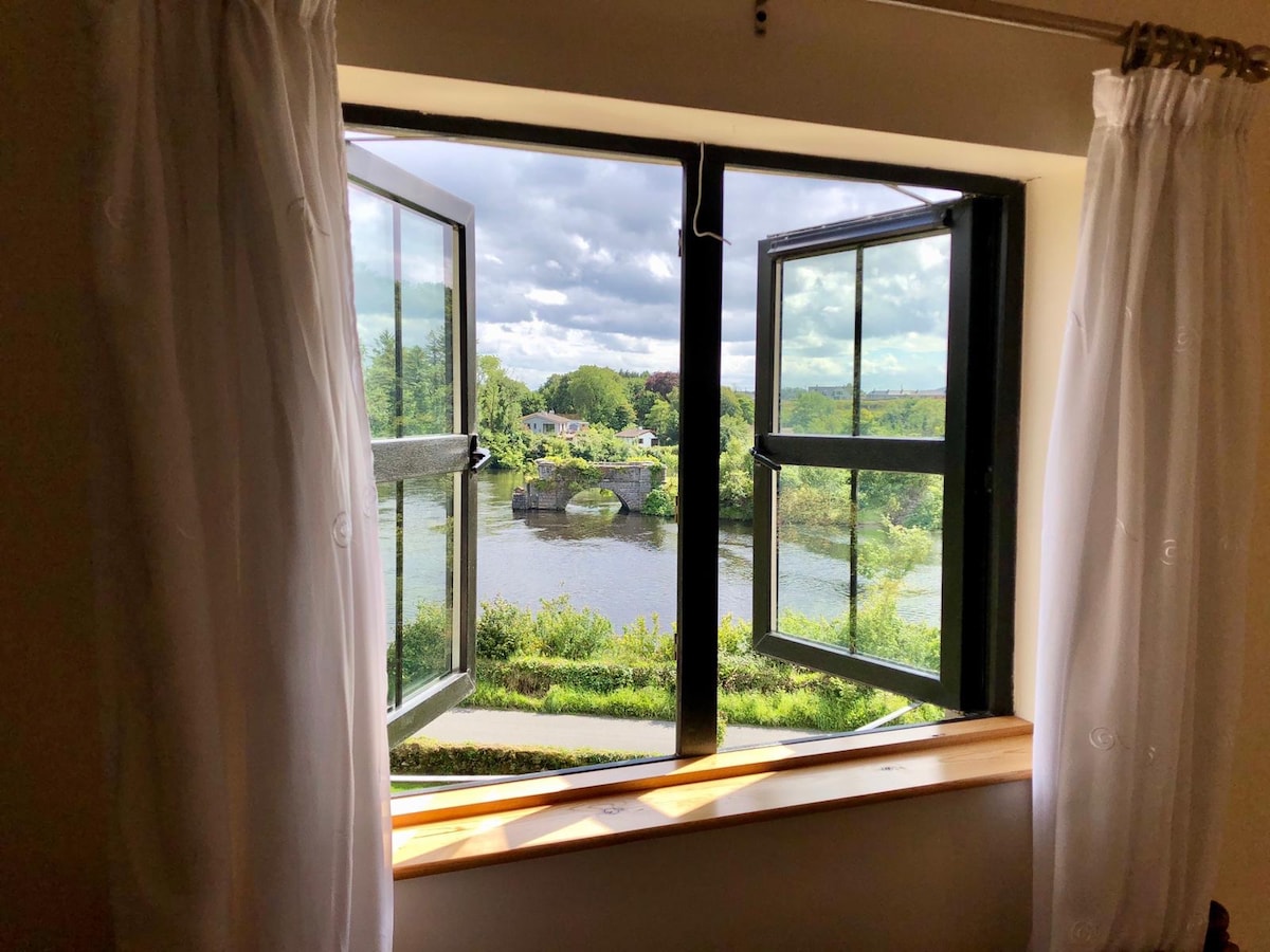 A window with dual casement designs is shown, framed by white curtains. The view reveals the tranquil River Erne and surrounding greenery, with clouds partially covering the sky. A wooden ledge sits below the window, creating an inviting space to enjoy the scenery.