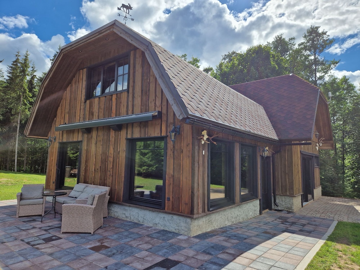 A two-story house features a rustic wooden exterior with a sloped roof and large windows. A stone patio extends in front, furnished with two comfortable chairs and a small table. Lush greenery surrounds the property, enhancing the natural setting.
