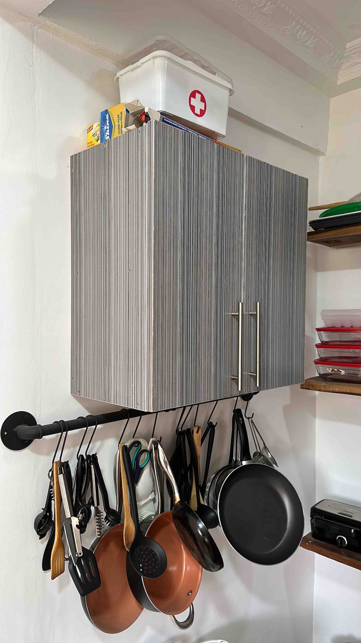 A kitchen cabinet with a sleek, grey-striped finish is mounted on the wall. It features two metal handles and is topped with a white box marked with a red cross. Below, a rod holds various cooking utensils, including metal, wooden, and copper pots and pans.