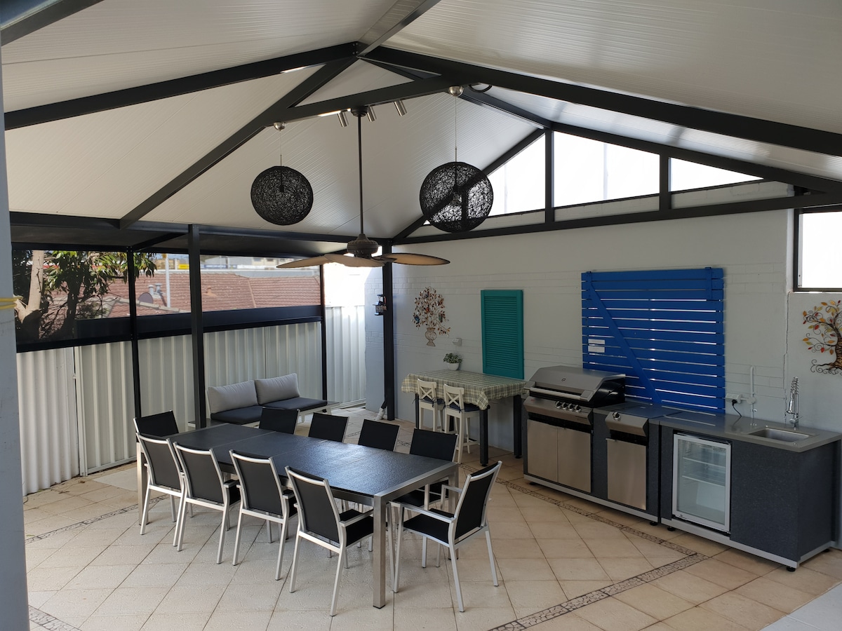 An outdoor living space is highlighted by a large granite dining table with ten chairs, complemented by a gas barbecue and a bar fridge. Natural light floods the area through high ceilings and windows, with decorative wall art adding character.