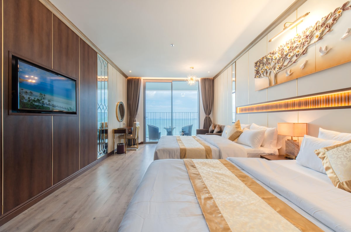 A spacious bedroom includes two queen beds adorned with gold-accented bed runners. Natural light floods the room through sliding glass doors, leading to a balcony with ocean views. A modern TV and tasteful wall decorations enhance the contemporary design.