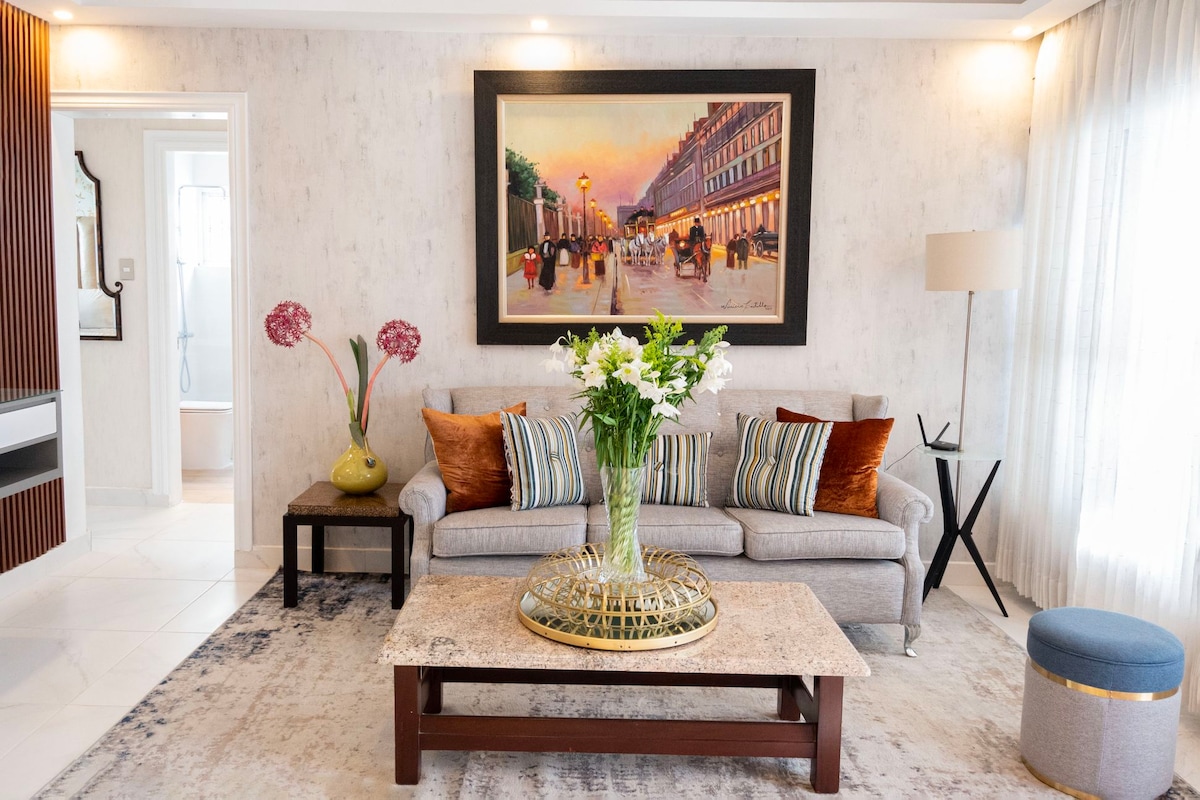 A comfortable sitting area features a light gray sofa adorned with colorful cushions, positioned across from a rectangular coffee table. A large floral arrangement occupies the center of the table. A vibrant cityscape artwork is displayed on the wall, complemented by soft lighting and sheer curtains.