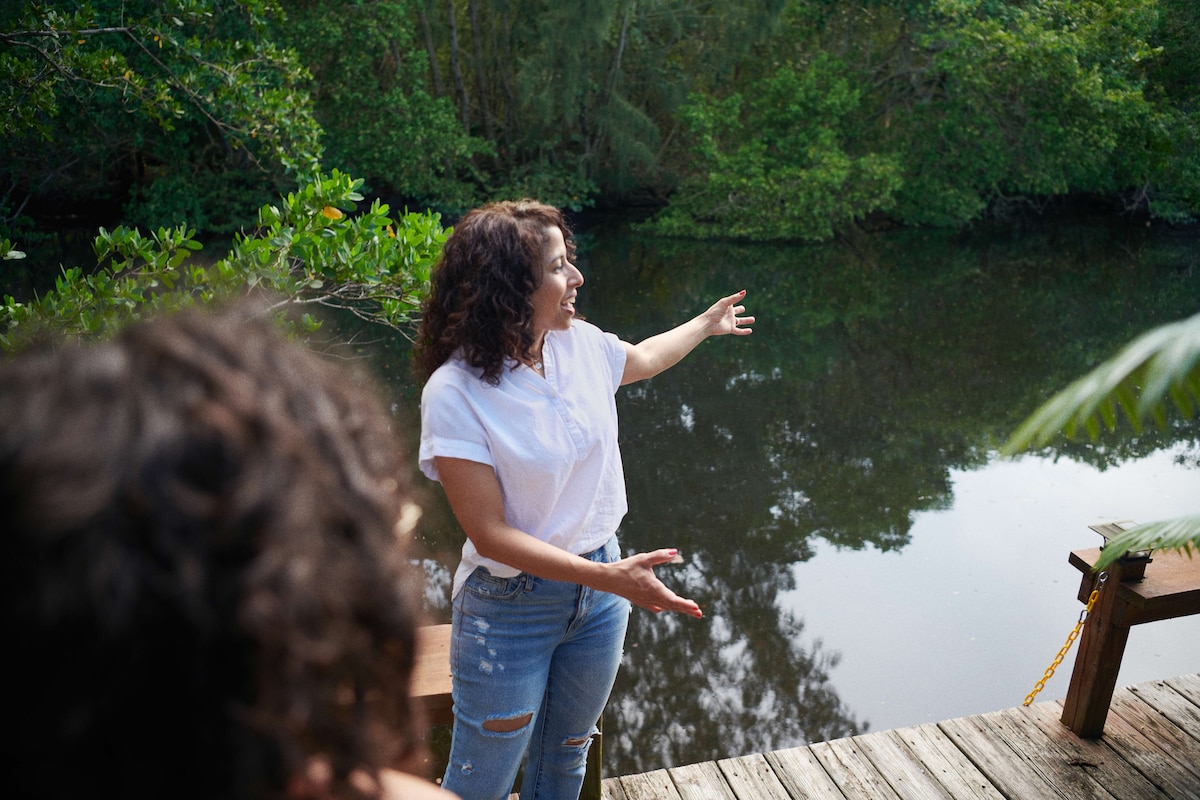 Host standing on a wooden deck and gesturing toward a calm river surrounded by lush greenery.