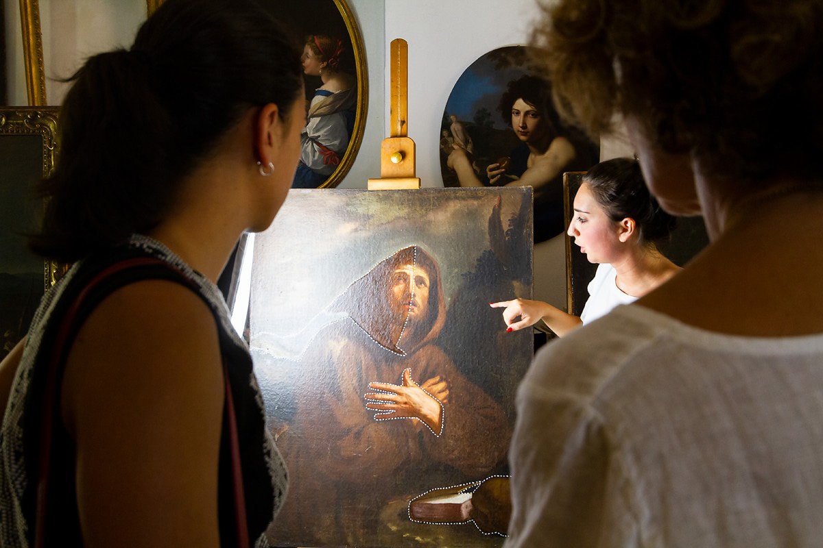 Host guiding guests through an art space while pointing at a historic painting on an easel.