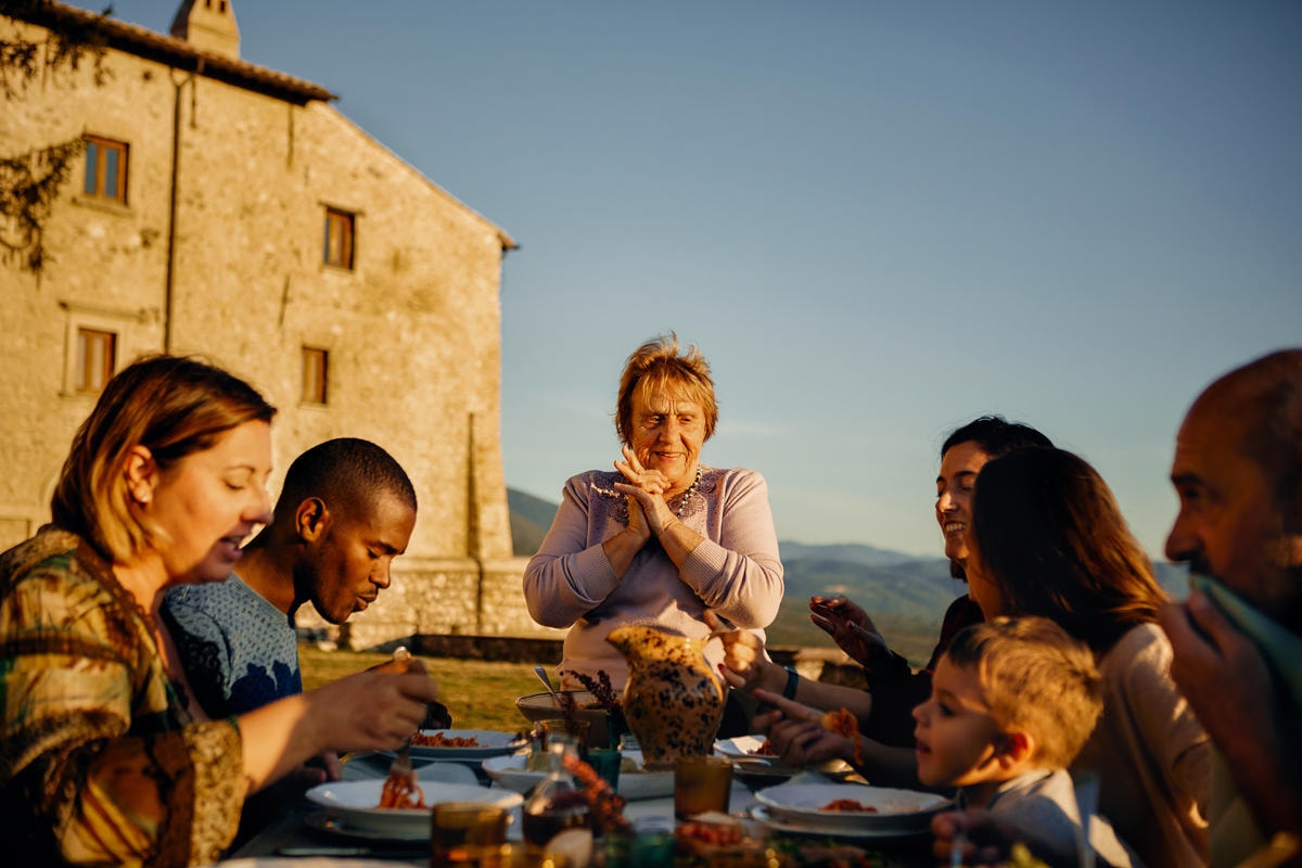 Host sharing an outdoor meal with guests at sunset beside a rustic countryside building.