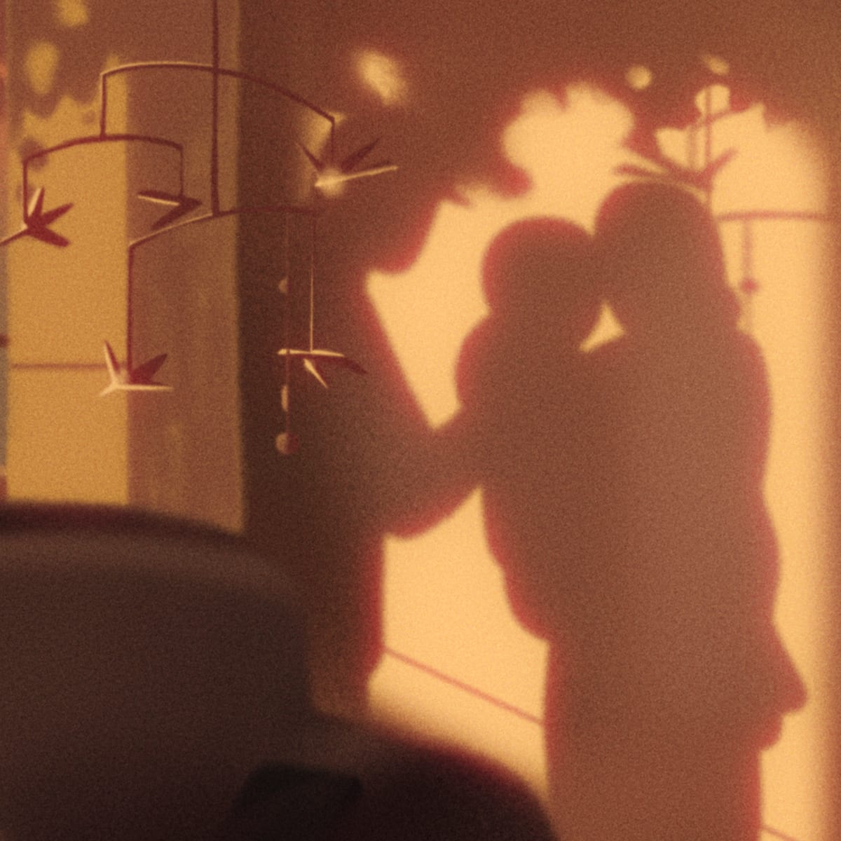 Illustrated silhouette of parents holding a child against a warm, glowing wall, with a paper crane mobile hanging nearby and toys and books visible on a shelf in the foreground.