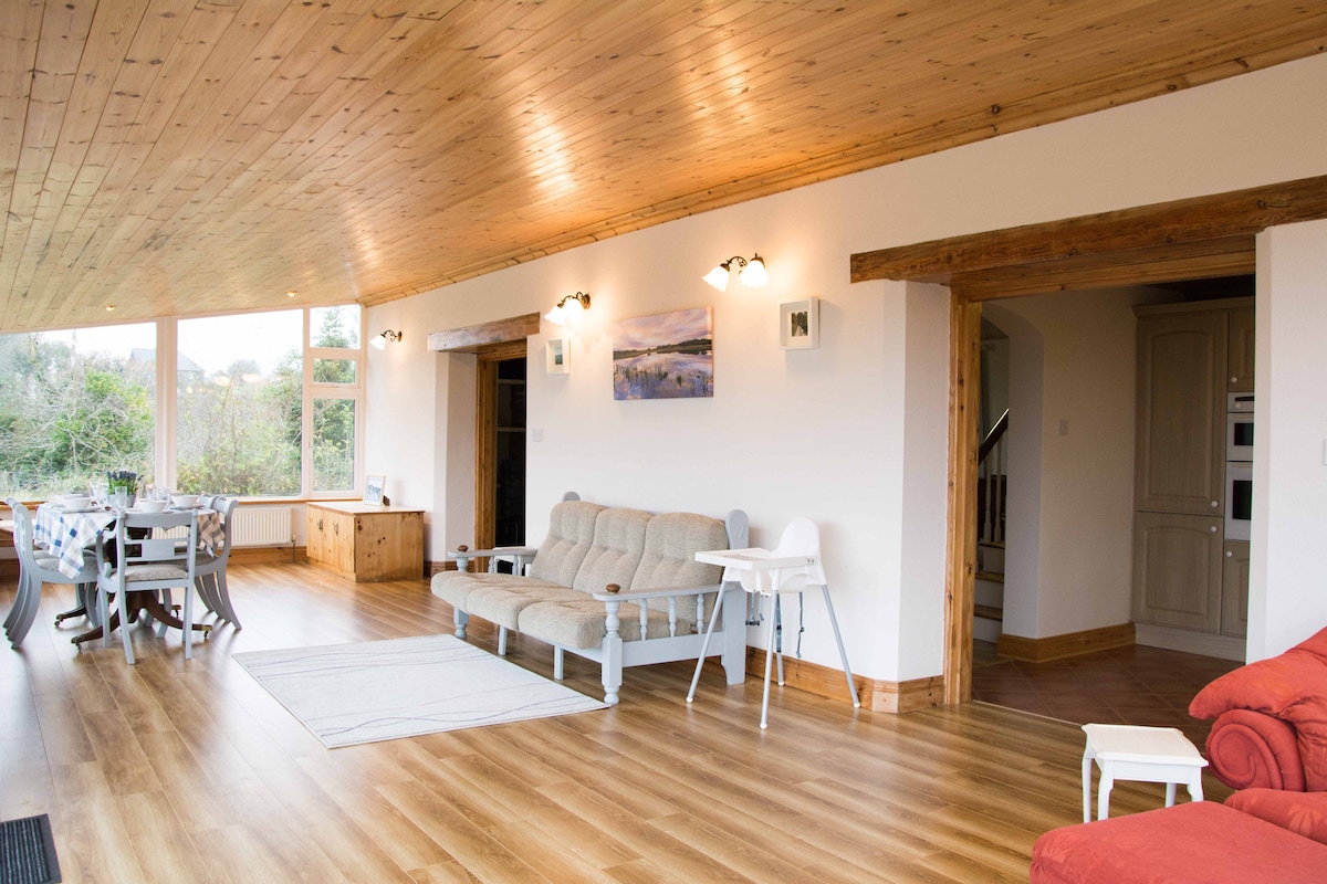 A spacious living area showcases wooden ceilings and large windows allowing natural light to fill the space. A comfortable sofa and a dining table with chairs are visible, complemented by light-colored flooring. A soft rug is placed centrally, adding to the inviting environment.
