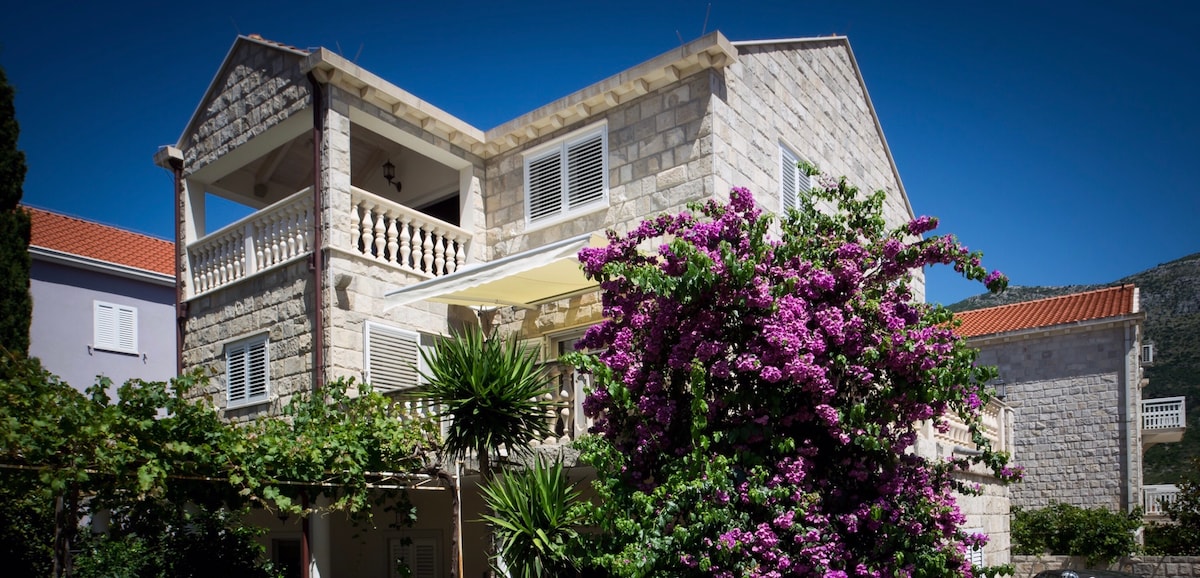 A traditional stone house is surrounded by vibrant greenery and blooming purple flowers. The structure features a spacious balcony with balustrades, offering a striking view of the landscape. Clear blue skies provide a bright backdrop to this welcoming and serene setting.