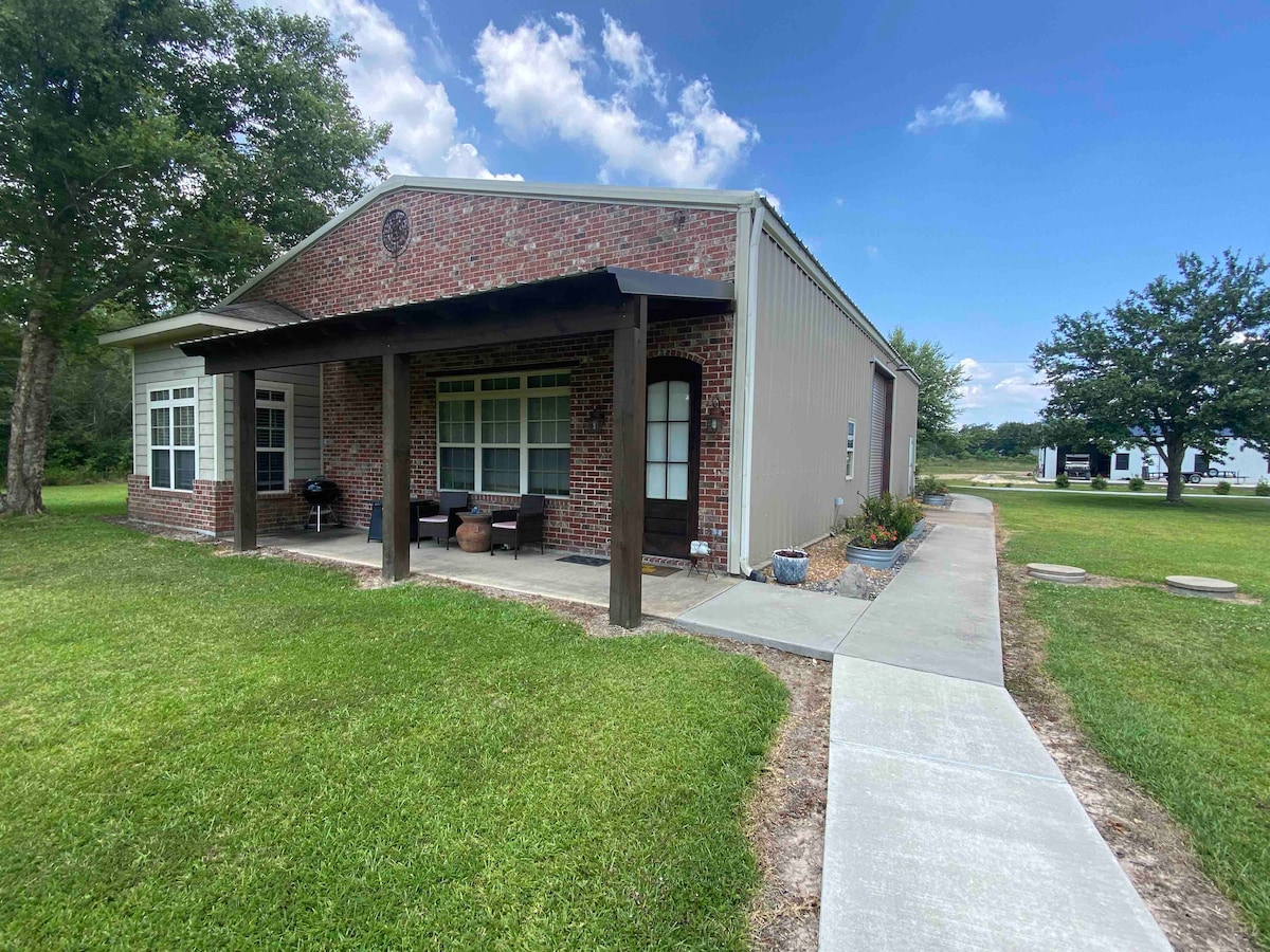 The exterior of the carriage house is presented, featuring a combination of brick and metal siding. A covered porch with seating is visible, alongside a paved walkway leading to the entrance. Lush green grass surrounds the home, with trees and open space in the background.