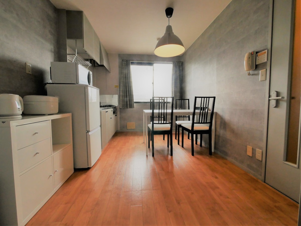 A modern kitchen area features a dining table with four black chairs. White cabinetry and appliances, including a microwave and refrigerator, are visible. Natural light enters through the window dressed with light curtains, highlighting the warm wooden floor.
