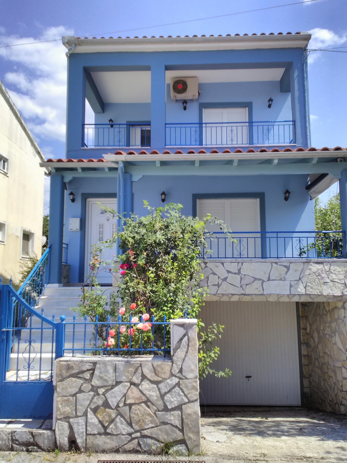 The exterior of a two-story house is presented, featuring a vibrant blue facade and white accents. The entrance is framed by steps leading to a covered porch, adorned with flowering plants. A garage is visible below, and light fixtures illuminate the space.