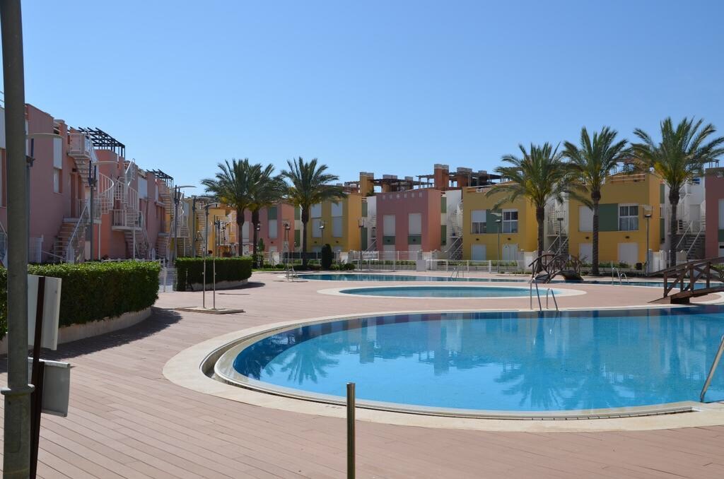 A community pool area is surrounded by well-maintained palm trees and colorful residential buildings. The pool features a circular design with clear blue water, and a wooden deck offers space for relaxation. Ample sunlight fills the scene under a clear blue sky.