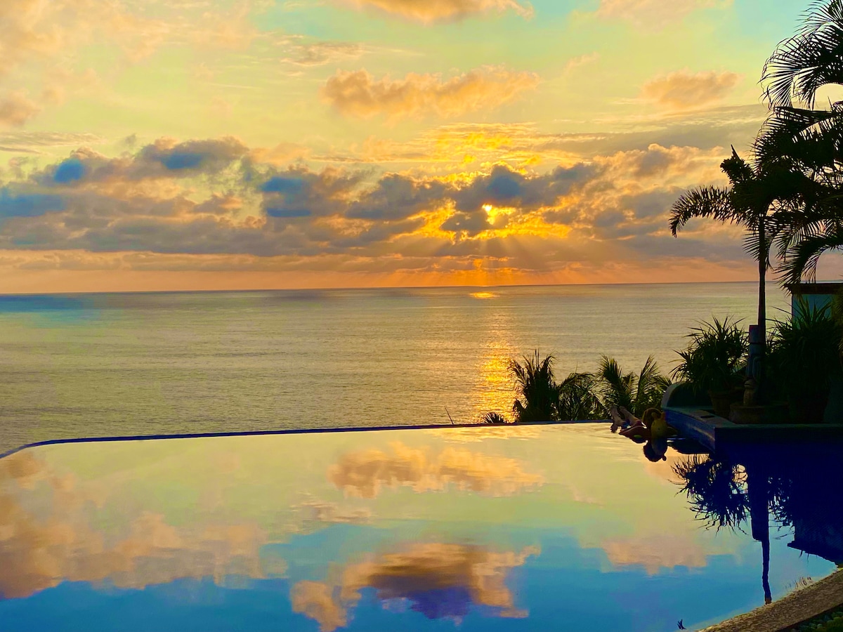An infinity pool reflects the vibrant colors of a sunset over the ocean. Silhouettes of lush tropical greenery frame the scene, enhancing the tranquil atmosphere. Gentle waves can be seen in the background, creating an inviting setting for relaxation.